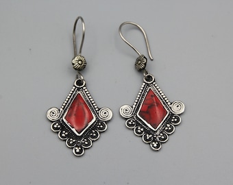 May include: A pair of silver-coloured drop earrings. Each earring has a red, diamond-shaped stone set in a decorative frame. The frame has a floral design at the bottom and a spiral design on each side. The earrings have a hook closure.