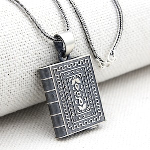 Puede incluir: Un collar de plata con un colgante en forma de libro cerrado. El libro tiene un diseño intrincado con un acabado negro y detalles plateados.