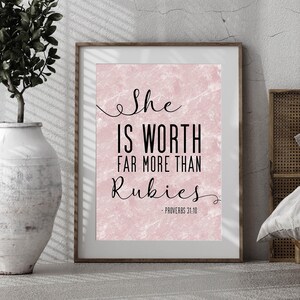 May include: A framed print with the text "She IS WORTH FAR MORE THAN Rubies - PROVERBS 31:10" in black script on a pink marble-textured background. The frame is a light brown color. A white vase with a plant is visible on the left.