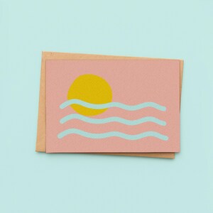 May include: A greeting card with a pink background featuring a yellow sun and light blue wavy lines, resembling ocean waves. The card is placed on top of a brown envelope, set against a light blue backdrop.