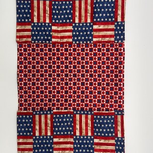 May include: A rectangular piece of fabric with a patchwork design. The top and bottom sections feature a pattern resembling the American flag, with blue fields of white stars and red and white stripes. The middle section has a red and blue star pattern.