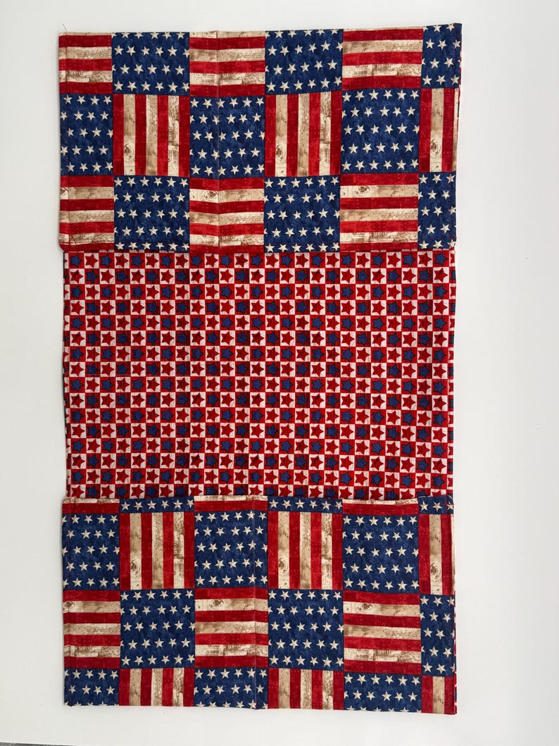 May include: A rectangular piece of fabric with a patchwork design. The top and bottom sections feature a pattern resembling the American flag, with blue fields of white stars and red and white stripes. The middle section has a red and blue star pattern.