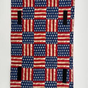 May include: A rectangular textile with a patchwork design of the American flag. The design includes blue squares with white stars and red and white striped sections. Four black straps are sewn onto the textile.