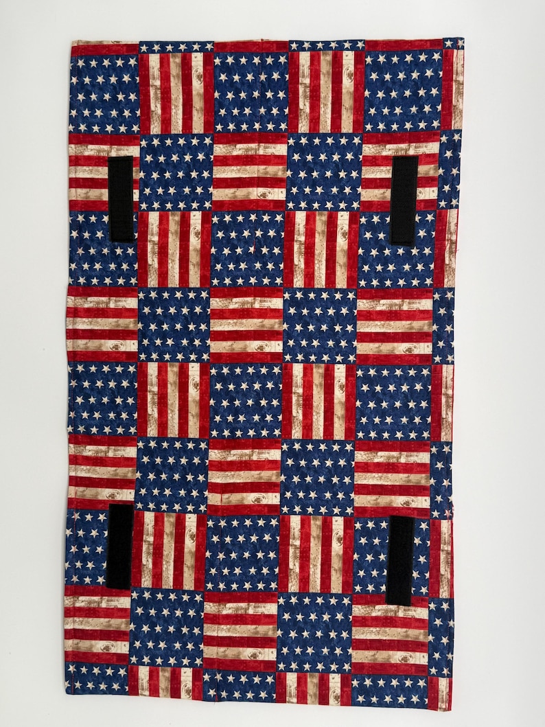 May include: A rectangular textile with a patchwork design of the American flag. The design includes blue squares with white stars and red and white striped sections. Four black straps are sewn onto the textile.