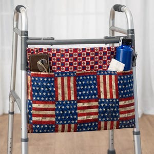 May include: A silver walker with a fabric organiser. The organiser has an American flag design and pockets containing a wallet, glasses, a blue water bottle, and tissues. The walker has black handles and is designed for mobility.