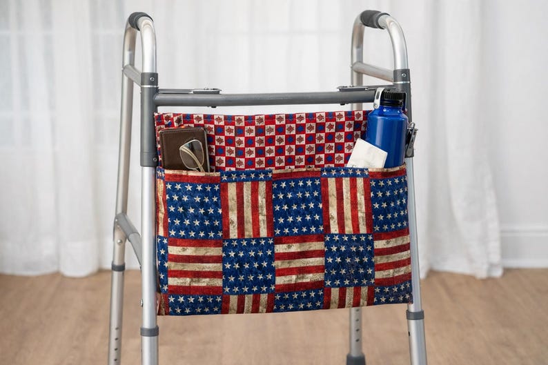 May include: A silver walker with a fabric organiser. The organiser has an American flag design and pockets containing a wallet, glasses, a blue water bottle, and tissues. The walker has black handles and is designed for mobility.