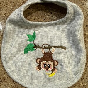 May include: A gray waffle-weave baby bib with a brown monkey embroidered on the front. The monkey is hanging from a brown branch with green leaves and holding a yellow banana. The bib has a rounded shape and a snap closure.