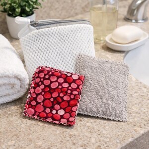Pode incluir: Uma cole&ccedil;&atilde;o de acess&oacute;rios de casa de banho numa bancada. Inclui um saco de malha branco com fecho cinzento, um quadrado de bolinhas vermelhas e rosa, um quadrado texturizado cinzento, um saboneteira com sab&atilde;o e um dispensador de sab&atilde;o transparente.