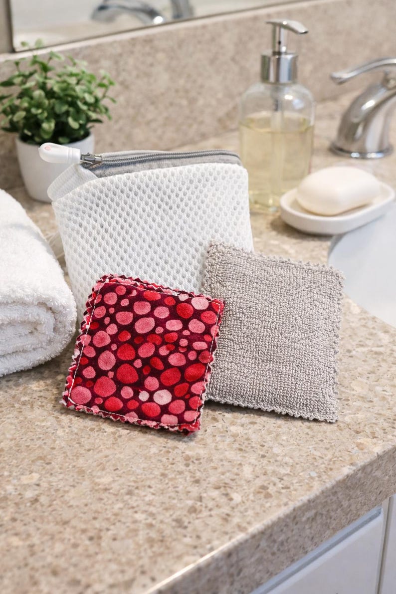 Pode incluir: Uma cole&ccedil;&atilde;o de acess&oacute;rios de casa de banho numa bancada. Inclui um saco de malha branco com fecho cinzento, um quadrado de bolinhas vermelhas e rosa, um quadrado texturizado cinzento, um saboneteira com sab&atilde;o e um dispensador de sab&atilde;o transparente.