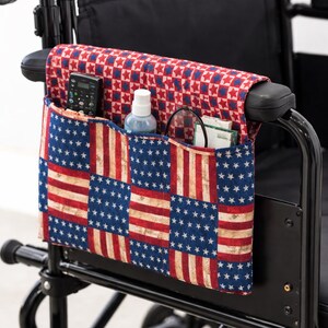 May include: Wheelchair armrest organiser with a patriotic American flag design. The fabric organiser has multiple pockets and a red, white, and blue colour scheme. It holds a remote control, spray bottle, and other items.