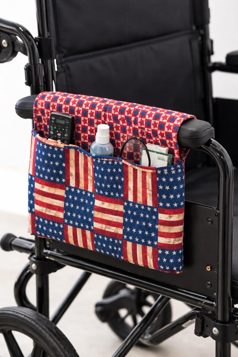 May include: Wheelchair armrest organiser with a patriotic American flag design. The fabric organiser has multiple pockets and a red, white, and blue colour scheme. It holds a remote control, spray bottle, and other items.