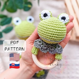 Może przedstawiać: Rattle zabawka w kształcie żaby wykonana na szydełku z drewnianym pierścieniem. Żaba jest zielona z białymi oczami i szarym szydełkowym kołnierzem. Zabawka jest wykonana ręcznie i dostępny jest wzór PDF.