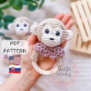 May include: Crochet monkey rattle toy with a wooden ring. The monkey is beige with brown eyes and a pink crocheted collar. The pattern is available in English and Slovak.