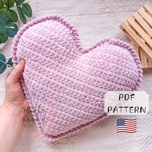 May include: A heart-shaped, light purple crochet pillow with a textured surface and a decorative border. The pillow is held in a hand. The image includes a small sign that says "PDF PATTERN" and an American flag.