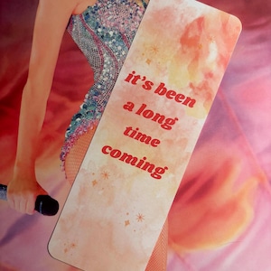 May include: A peach-colored bookmark with a watercolor background and gold glitter accents. The text "it's been a long time coming" is printed in red.