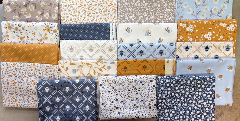 May include: A collection of folded fabric squares featuring various patterns including floral prints, small white daisies, repeating bee motifs, polka dots, and leafy vines in a color palette of navy blue, mustard yellow, light blue, white, and beige.