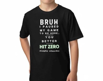 May include: Black t-shirt with white text that reads "BRUH I PAUSED MY GAME TO BE HERE... YOU BETTER HIT ZERO." The hashtags #CompDay and #CheerBro are below the text. The shirt is being worn by a person.