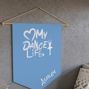 May include: A light blue pennant banner with the text "My Dance Life" in white, along with a heart and a ballerina silhouette. The name "Ashley" is written below. The banner is suspended from a wooden rod.
