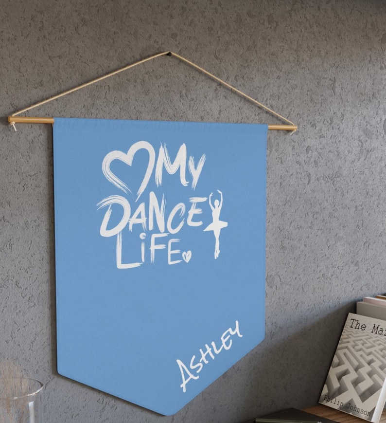May include: A light blue pennant banner with the text "My Dance Life" in white, along with a heart and a ballerina silhouette. The name "Ashley" is written below. The banner is suspended from a wooden rod.