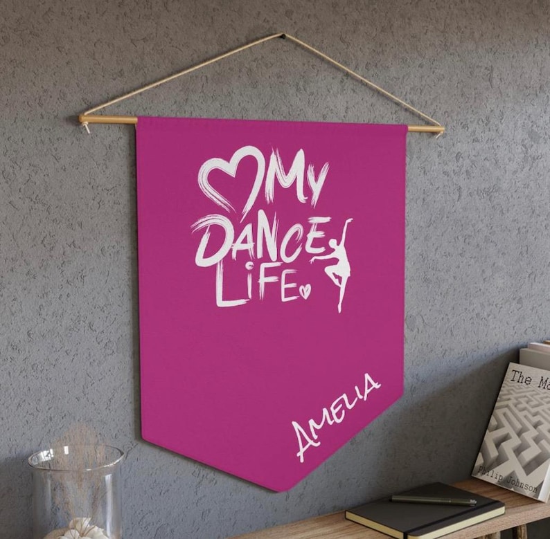 May include: A magenta pennant banner with the text "My Dance Life" in white script, featuring a heart and a dancer silhouette. The name "Amelia" is written below. The banner is suspended from a wooden rod with a string.