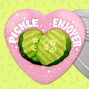 May include: A heart-shaped pin with a pink background and the words "Pickle Enjoyer" in white. The pin features two green pickle slices. The back of the pin is silver with a pin clasp.