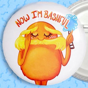 Puede incluir: Un pin de botón blanco con una ilustración de dibujos animados de un personaje naranja con cabello amarillo y bigote. El texto "NOW I'M BASHFUL" está escrito en naranja. El personaje sostiene un objeto azul.
