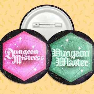 May include: Two 1-inch diameter button pins with a black background and a glittery, iridescent, multi-colored, 20-sided die design. The die is pink on the left pin and green on the right pin. The text "Dungeon Mistress" is on the left pin and "Dungeon Master" is on the right pin.