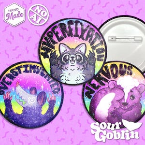 Puede incluir: Tres chapas redondas con alfiler e ilustraciones coloridas. Una muestra un mapache con el texto HYPERFIXATION, otra una zarigüeya con OVERSTIMULATED y otra un oso con NERVOUS. Incluye pequeños iconos blancos de Hand Made y No AI.