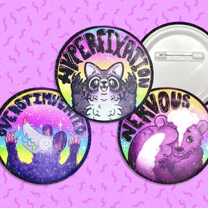 Op de afbeelding: Drie ronde knopen met kleurrijke achtergronden en glitter. De knopen tonen cartoondieren met tekst die "Overstimulated", "Hyperfixation" en "Nervous" zegt.