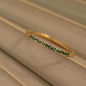 Puede incluir: Un anillo fino de color dorado con una fila de pequeñas piedras preciosas de color verde esmeralda. El anillo se presenta sobre una superficie texturizada de tonos neutros. El diseño es simple y elegante, mostrando el estilo minimalista y clásico del anillo.