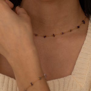 May include: A delicate gold necklace and bracelet set. The necklace and bracelet feature small, round gold beads and irregularly shaped purple gemstones. The jewelry is displayed against a cream-colored background, highlighting the gold and purple color scheme.