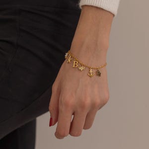 May include: Gold-colored charm bracelet with multiple charms, including letters, a flower, an anchor, and a zodiac symbol. The bracelet is worn on a wrist, showcasing the delicate chain and detailed charms.
