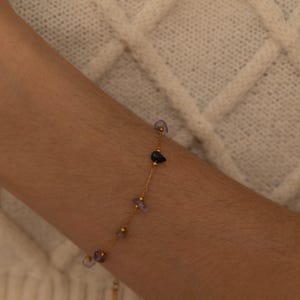 May include: A delicate gold bracelet featuring small, irregularly shaped gemstones in shades of purple, blue, and gold. The bracelet is strung on a fine gold chain, creating a minimalist and elegant design. The gemstones are spaced along the chain.