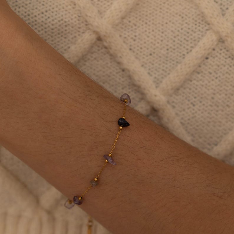 May include: A delicate gold bracelet featuring small, irregularly shaped gemstones in shades of purple, blue, and gold. The bracelet is strung on a fine gold chain, creating a minimalist and elegant design. The gemstones are spaced along the chain.