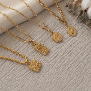 May include: A collection of four gold-toned necklaces featuring distinct chain styles and pendants. The pendants include a square textured charm, a rectangular floral plaque, a hexagonal rose motif, and an oval floral medallion on a curb chain.