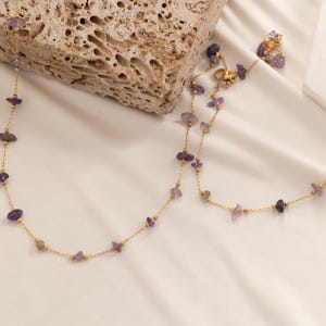 May include: A gold-colored necklace with small, irregularly shaped purple gemstones. The necklace is displayed on a white surface, with a textured stone block in the background. The gemstones are spaced along the delicate chain.