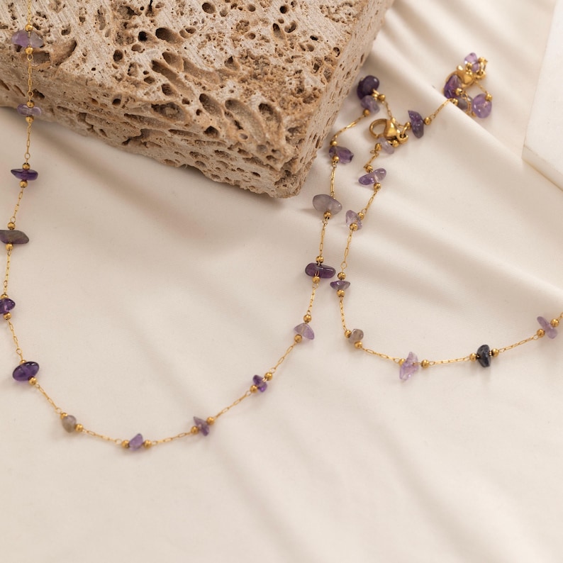 May include: A gold-colored necklace with small, irregularly shaped purple gemstones. The necklace is displayed on a white surface, with a textured stone block in the background. The gemstones are spaced along the delicate chain.