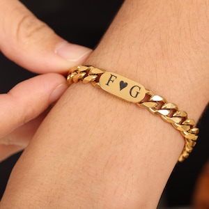 May include: Gold-colored curb chain bracelet with a rectangular plate. The plate is engraved with the letters "F", a heart symbol, and "G". The bracelet is a bright, shiny gold color.
