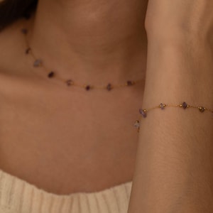 May include: A delicate gold necklace and bracelet set. The jewelry features small, irregularly shaped purple gemstones interspersed with tiny gold beads. The necklace is around the neck, and the bracelet is on the wrist.