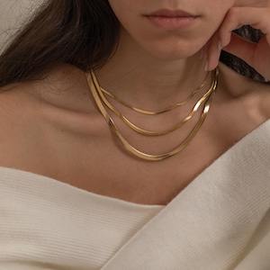 May include: Three gold herringbone chain necklaces layered on a person's neck. The necklaces vary in thickness, with the thickest at the bottom. The person is wearing a white off-the-shoulder sweater.