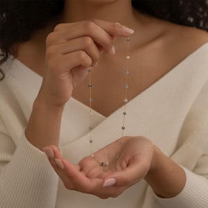 May include: A fine gold necklace adorned with small, round, blue and white gemstones. The necklace is displayed against a white sweater, highlighting the jewellery's design and colour contrast. A simple, elegant accessory.