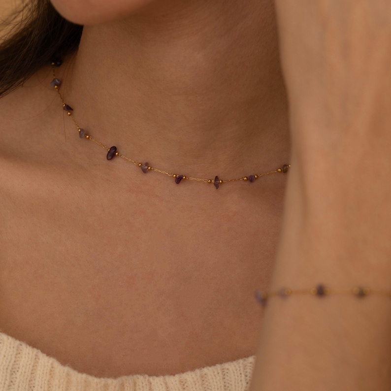 May include: Delicate gold necklace and bracelet set. The necklace features small, irregular purple gemstones and gold beads strung along a fine gold chain. The bracelet mirrors the necklace design. The jewelry is displayed on a person.