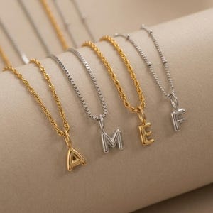 May include: A collection of gold and silver-toned initial necklaces. The necklaces feature different chain styles, with each pendant displaying a letter of the alphabet. The letters are in a simple, sans-serif font.