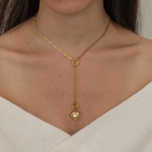 May include: A gold-toned lariat necklace featuring a mixed-link chain design. The upper portion consists of a long-link chain on one side and a smaller cable chain on the other, meeting at a central ring. A vertical drop chain ends in a heart charm.