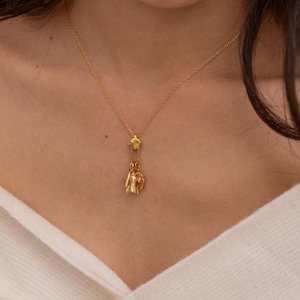 May include: Gold-colored necklace with a pendant featuring a small, detailed figure and a pink heart. The necklace is worn around the neck, with the pendant hanging at the chest. The chain and pendant have a shiny finish.
