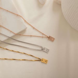 May include: Three necklaces with rectangular pendants featuring engraved letters. The top necklace is rose gold with a beaded chain and letter S. The middle is silver with a box chain and letter G. The bottom is gold with a twisted chain and letter A.