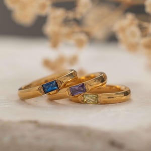 May include: Three gold rings with rectangular gemstones in blue, purple, and green. The rings are set on a light-colored surface, with a blurred background of dried flowers. The rings are likely for jewelry.