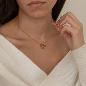 Puede incluir: Un collar de tono dorado con una cadena delicada de pequeñas cuentas y un colgante central formado por dos anillos entrelazados, uno de oro liso y otro más pequeño decorado con diminutas piedras de color violeta claro.