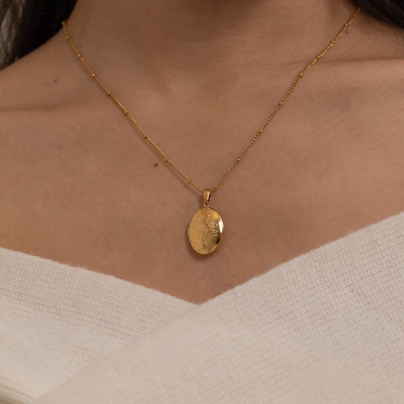 May include: A gold-colored oval locket necklace with a floral design. The pendant hangs from a delicate chain with small, evenly spaced beads. The necklace is worn against a white sweater.