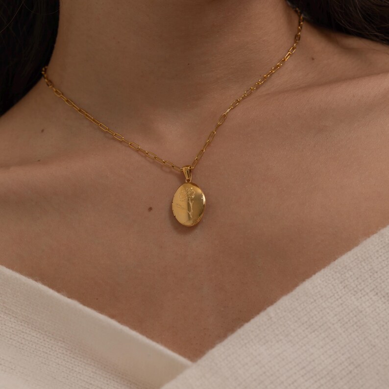 May include: A gold-colored necklace with a pendant. The pendant is an oval locket with a detailed design. The necklace chain is a delicate link style. The jewelry is worn around the neck.